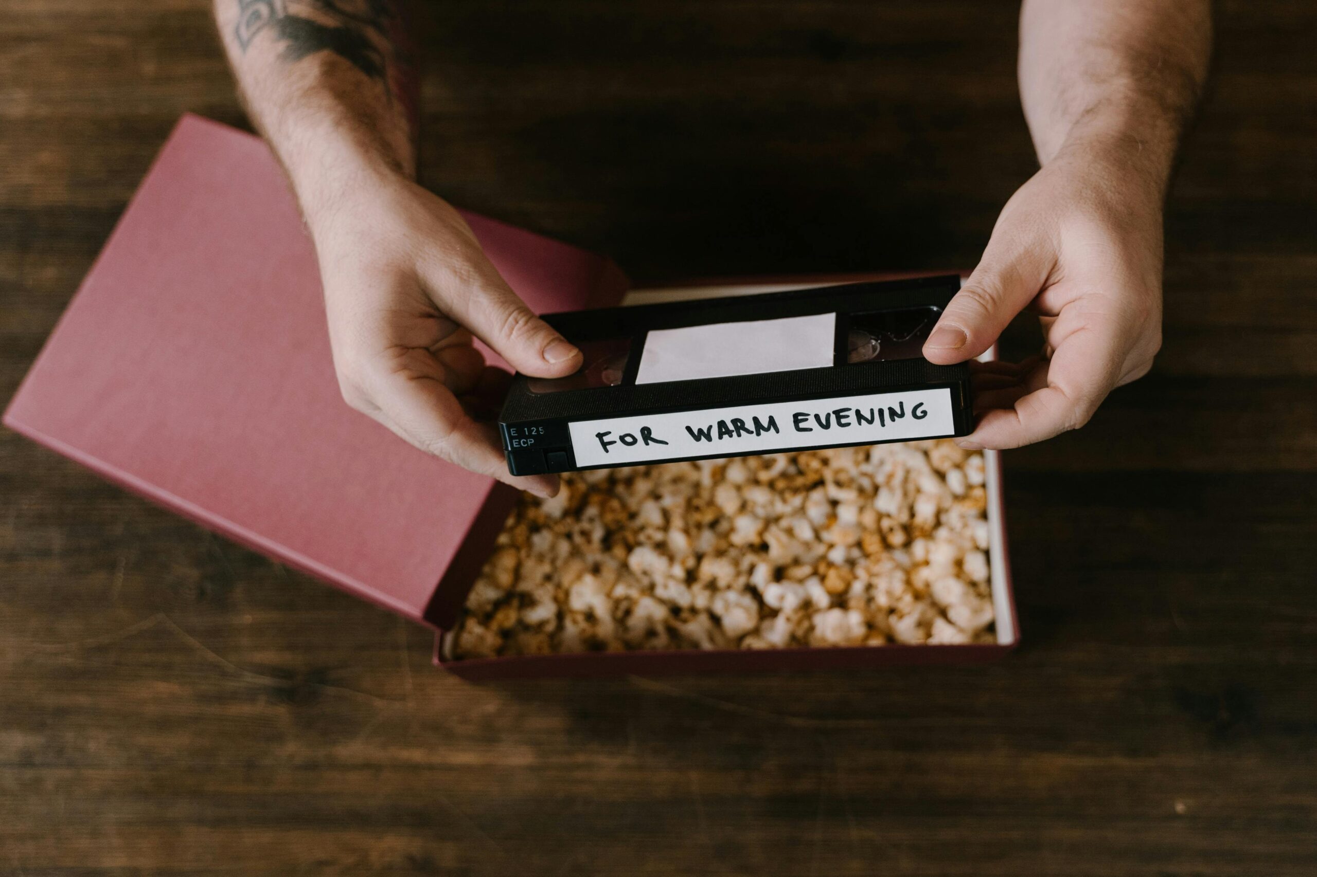 Hands holding a vintage VHS tape over a box of popcorn on a wooden surface.